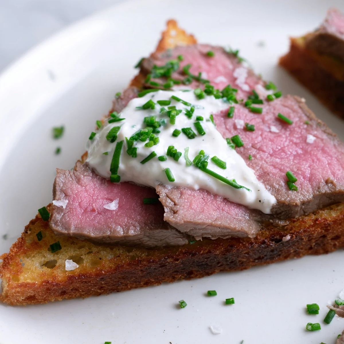 Golden-brown Beef Toast Points topped with tender seared beef slices, creamy horseradish sauce, and fresh chives on a rustic wooden board.