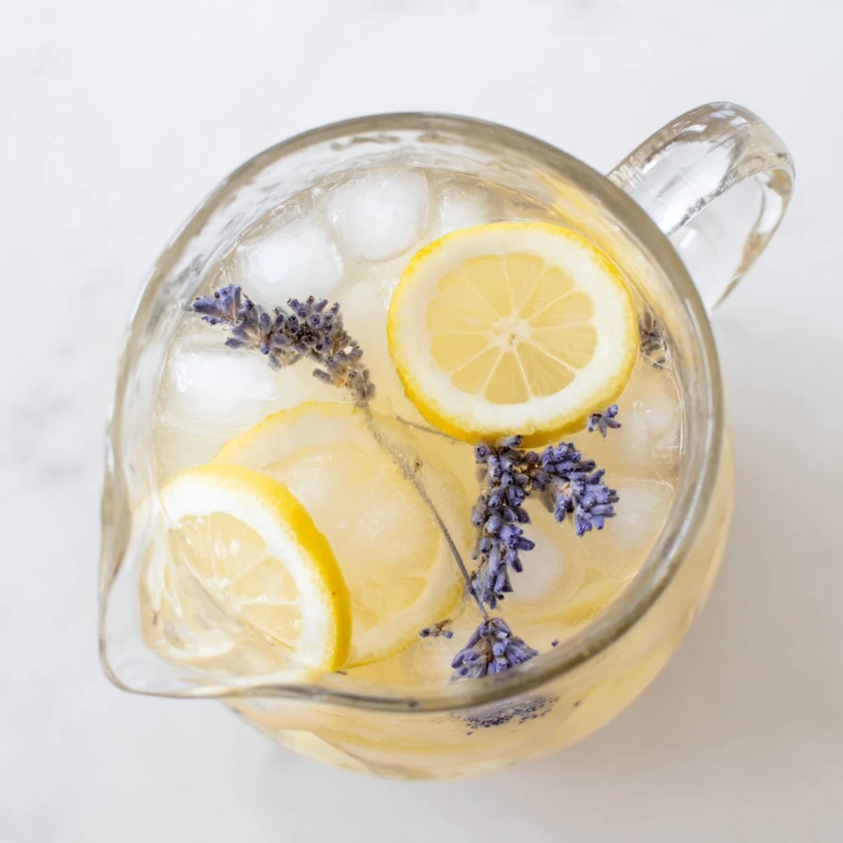 Glass pitcher of lavender lemonade garnished with fresh lemon slices and purple lavender sprigs on ice