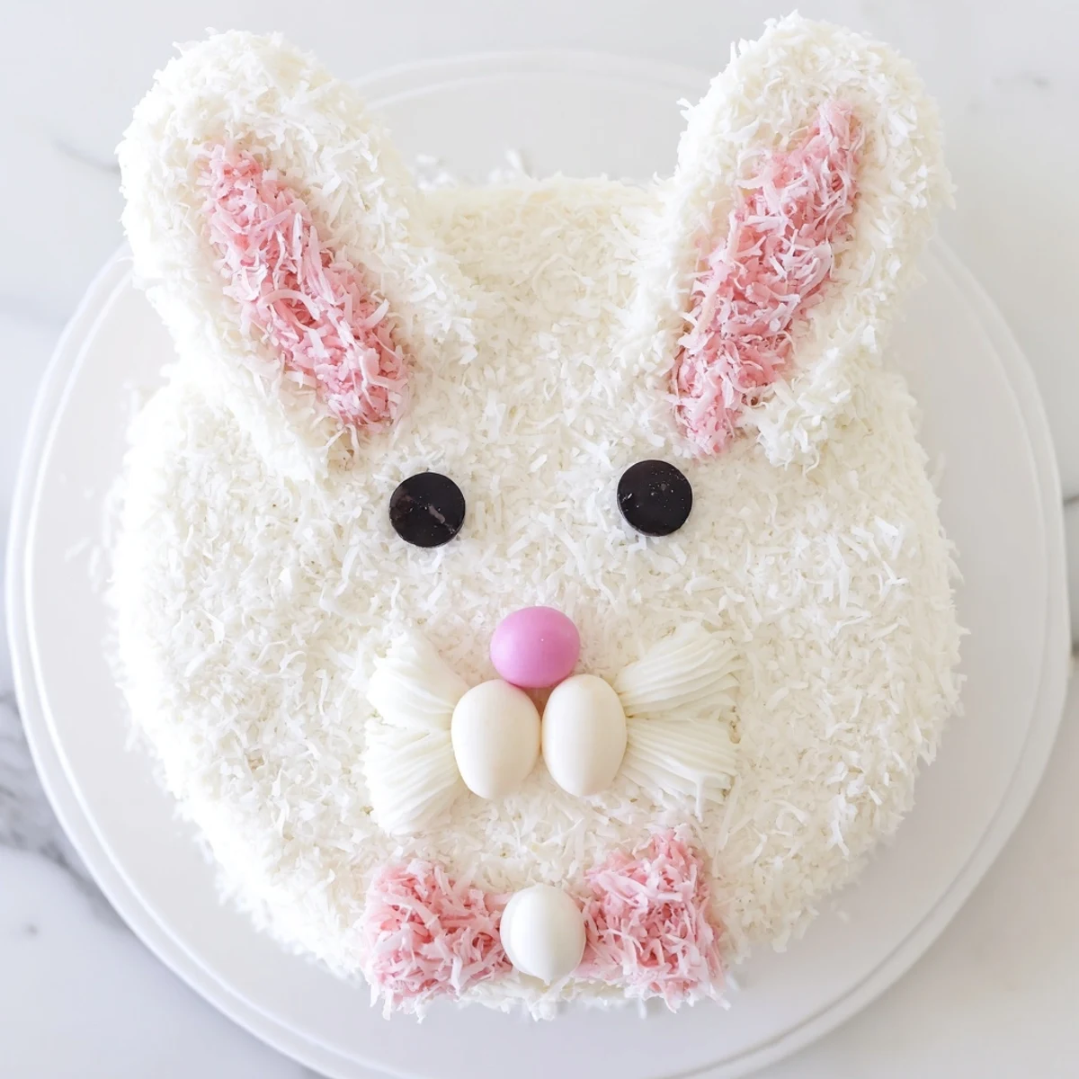 Adorable Bunny Cake covered in fluffy coconut frosting with pink fondant ears and jellybean features