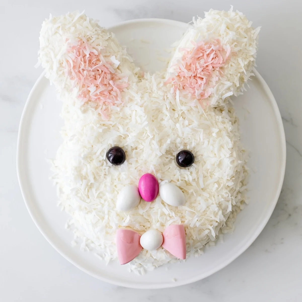 Whimsical vanilla Bunny Cake shaped with round face, bow tie body, and coconut-covered frosting details