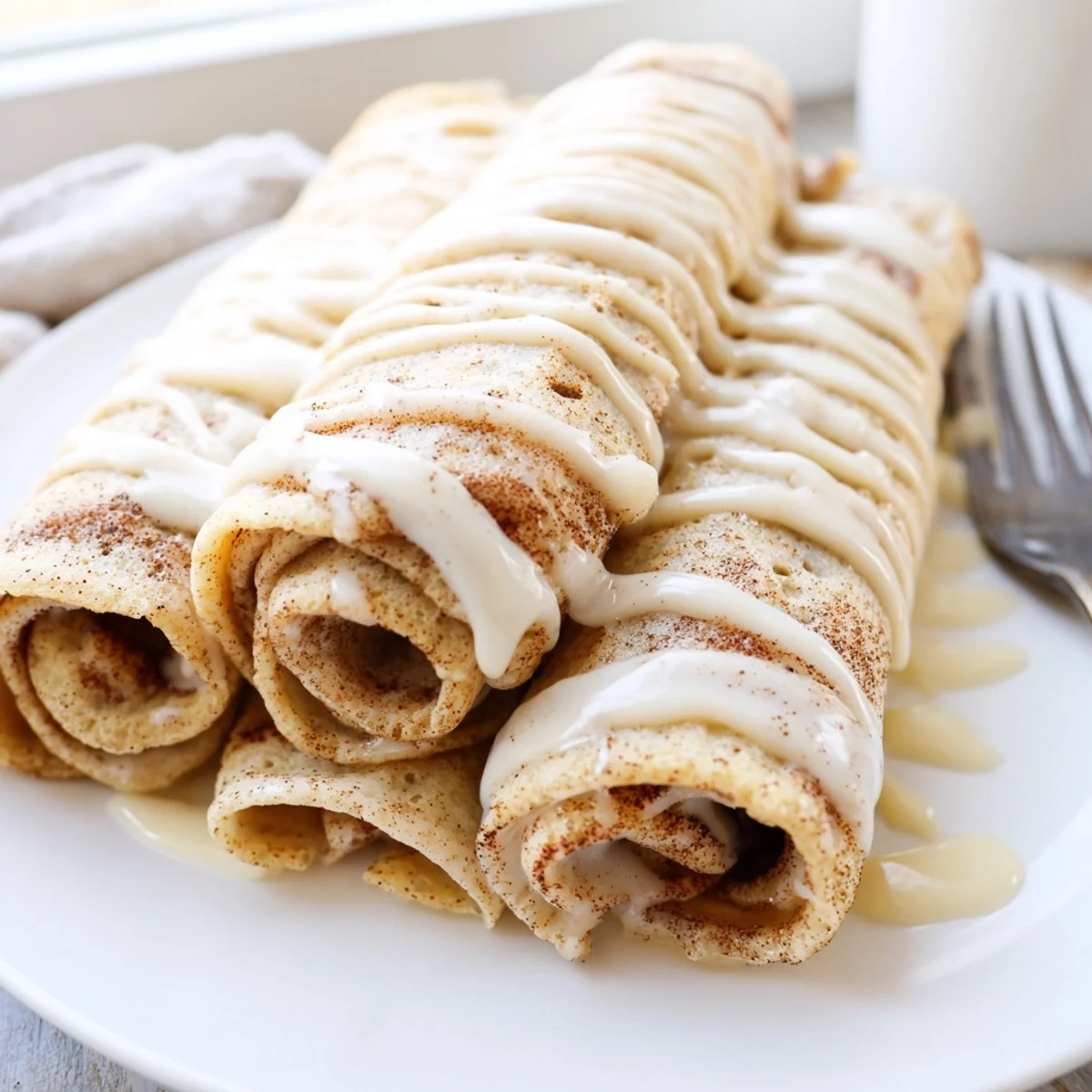 Golden cinnamon roll protein crepes drizzled with creamy vanilla glaze on a white plate